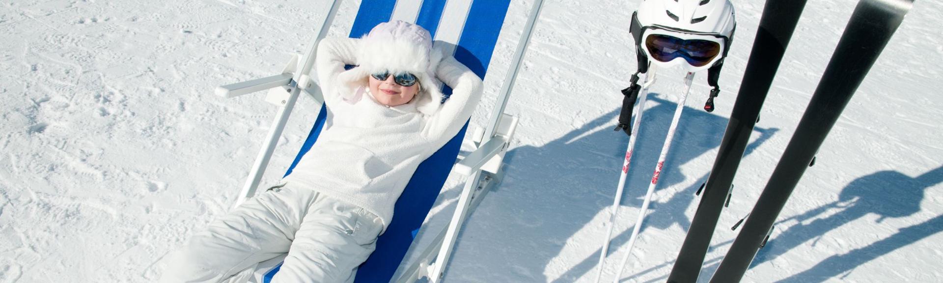 A serene winter scene on a snowy slope featuring a young child relaxing in a blue and white striped deck chair. Skis, ski poles, and a helmet with goggles stand upright in the snow nearby, with bright sunlight casting long shadows across the white snow.