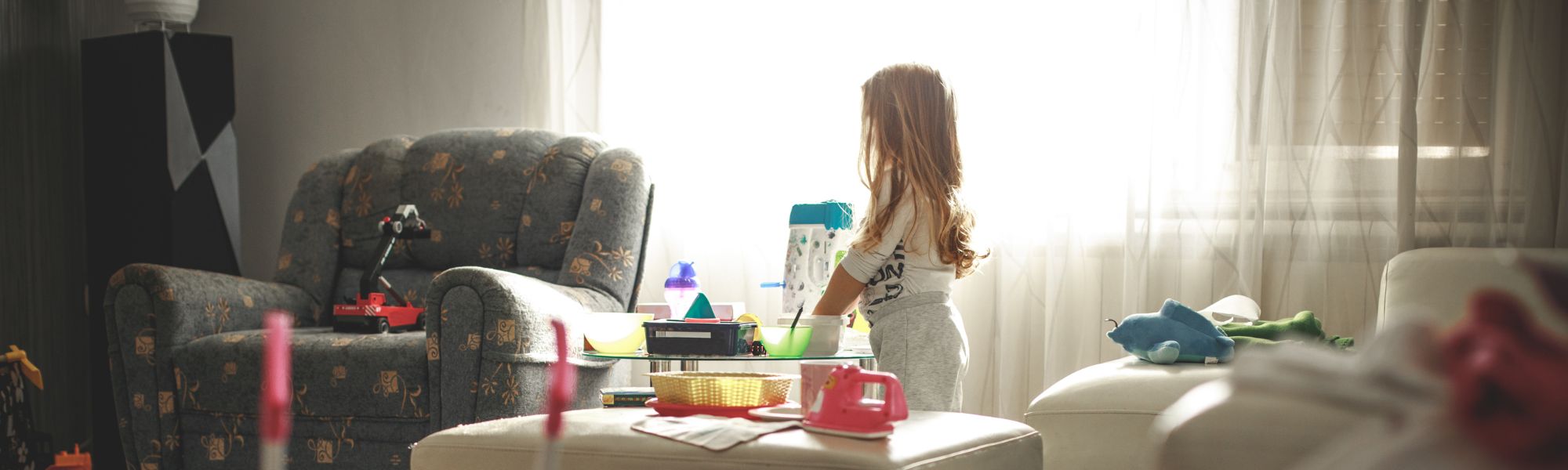 Young child standing among scattered toys in a messy living room, colourful toys covering the coffee table and armchair