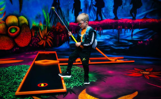 Young boy playing glow-in-the-dark mini-golf in a vibrant, neon-lit indoor setting. He holds a yellow putter, focusing on a glowing yellow golf ball on a black putting green with orange borders. The background features colorful, fluorescent murals of abstract plants, glowing landscapes, and silhouettes of hikers, creating an adventurous and immersive atmosphere.