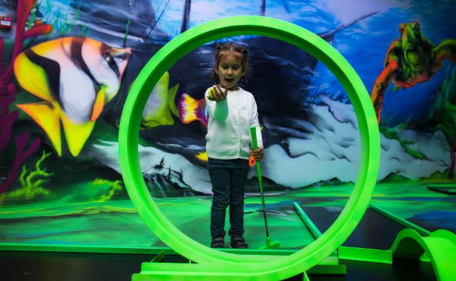 Young child playing indoor mini golf, standing inside a glowing green circular obstacle in a colorful underwater-themed course.