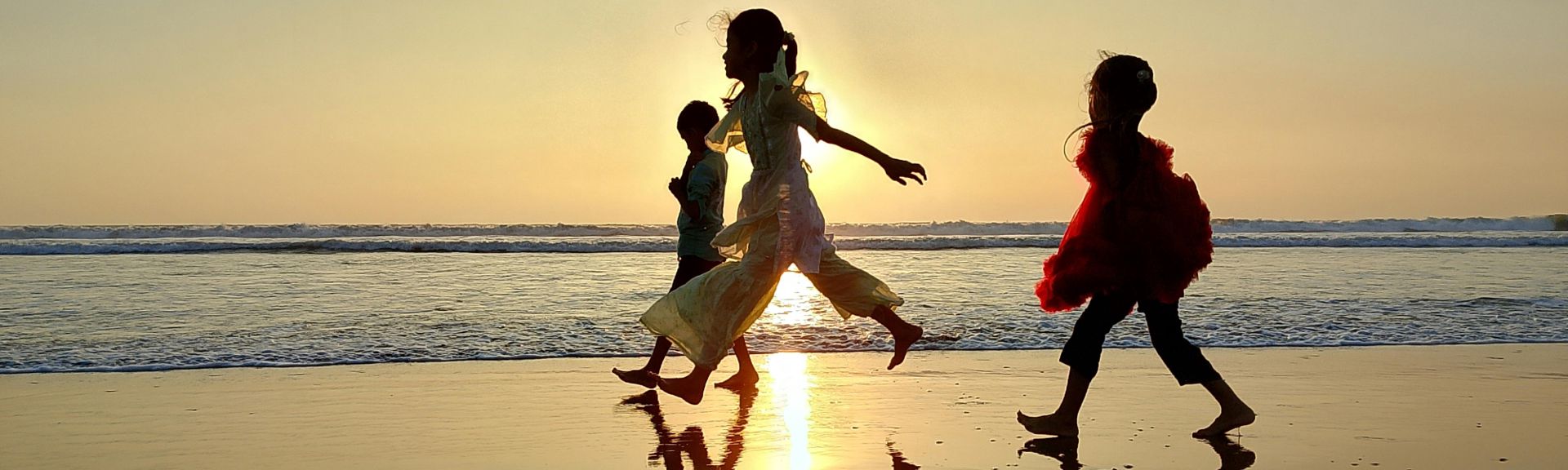 Children in flowing dresses running along the shoreline at sunset, silhouetted against the golden sky over the sea