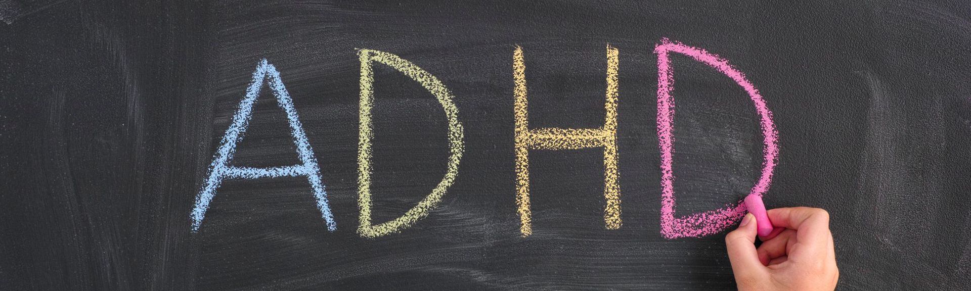 Colorful chalk letters spelling 'ADHD' on a dark chalkboard, with a hand holding pink chalk completing the final letter, symbolizing education and awareness about ADHD.