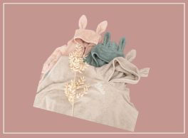 Three folded hooded baby towels with bunny ears, arranged diagonally on a rosy-pink background. The top left towel is light pink, the middle is muted teal, and the bottom right is beige with a subtle white emblem. A delicate dried floral element is placed on the beige towel, adding a soft, artisanal touch.