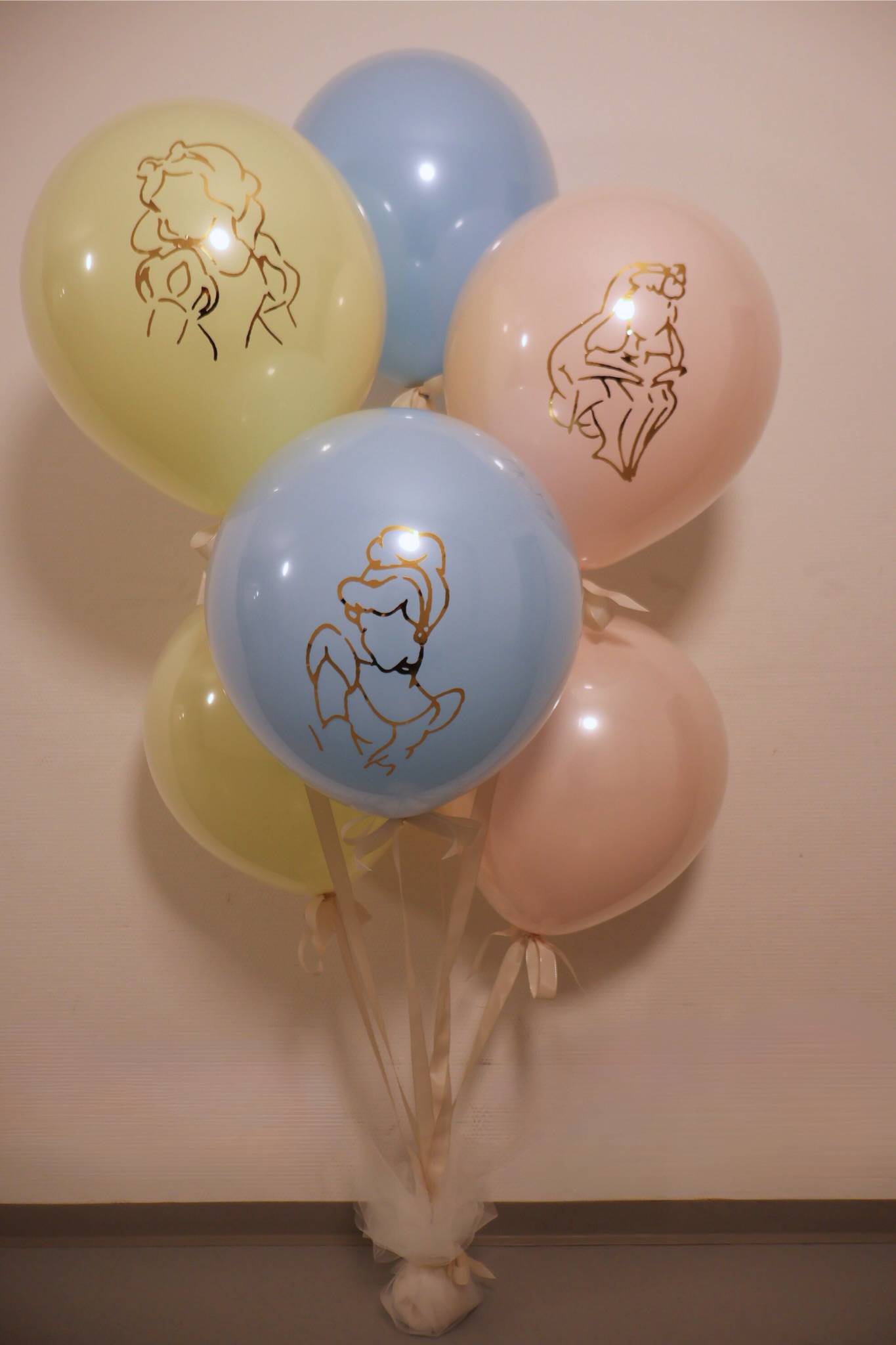 A cluster of six pastel-colored balloons featuring golden outlines of Disney princesses, likely Cinderella, tied together with light-colored ribbons and anchored to a soft, decorative white base. The balloons are in shades of light yellow, blue, and pink, arranged in an oval shape against a light-textured wall background. The overall scene exudes a whimsical and celebratory vibe.