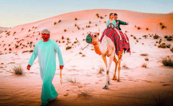 A vibrant desert scene featuring a man in traditional attire leading a camel carrying two smiling riders across warm, reddish-orange sand dunes. The camel is adorned with a colorful blanket, and the soft lighting suggests a serene sunrise or sunset.