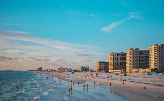 A lively beach scene during golden hour, with numerous people enjoying the light-colored sand and teal-blue ocean waves. Modern multi-story buildings line the coastline, while the warm sunlight and clear sky create a vibrant, inviting atmosphere.