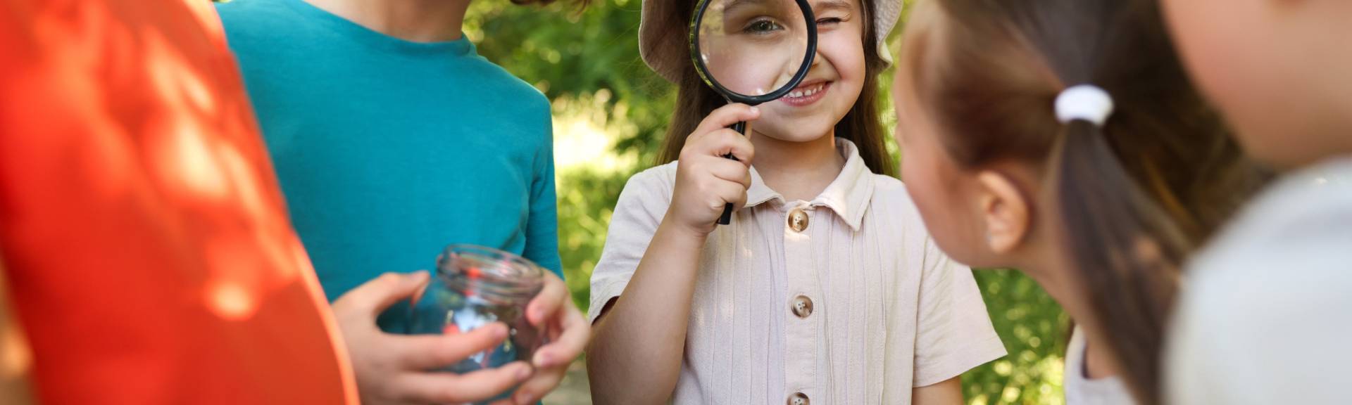 Smiling girl in a sun hat peers through a magnifying glass outdoors while friends hold a glass jar for their nature finds.