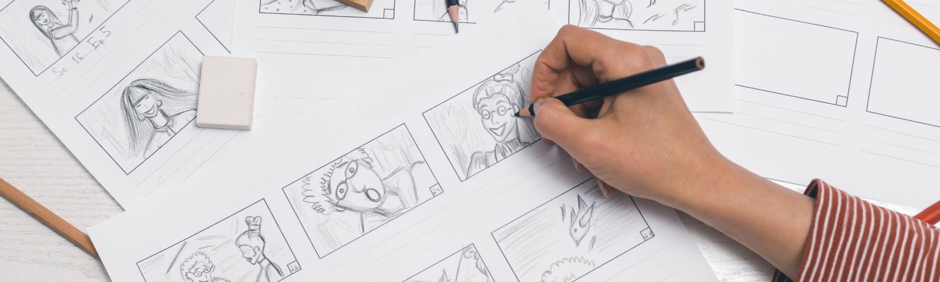 A close-up of a hand drawing storyboard panels on white paper spread across a light wooden table. The hand, wearing a reddish-brown and white striped sleeve, holds a black pencil sketching a character's face in one panel. Other panels feature pencil-drawn characters and scenes, with some blank frames visible. Pencils, an eraser, and a small wooden block are scattered around, emphasizing the creative process of visual storytelling.