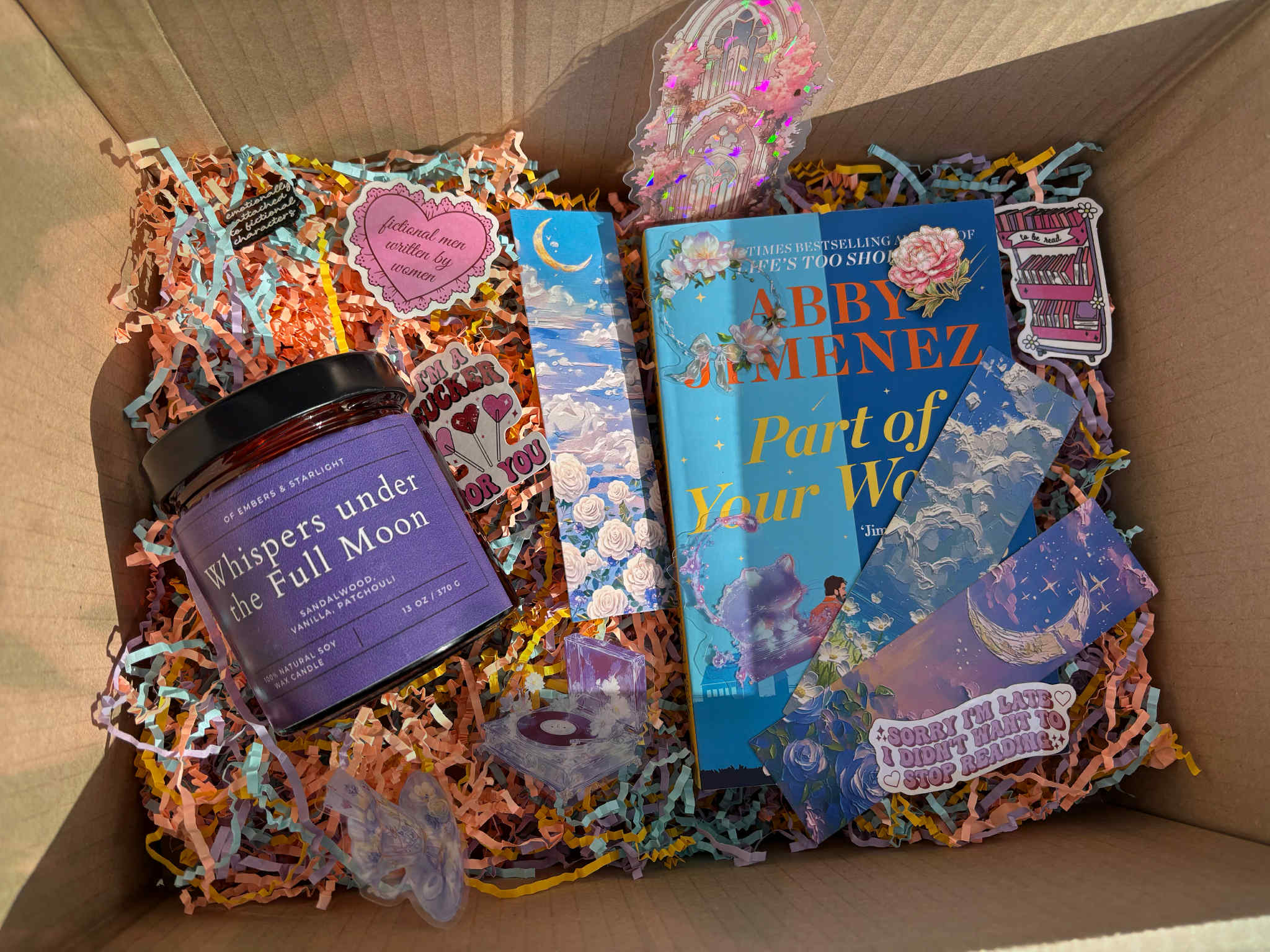 Book-themed gift box containing the novel Part of Your World by Abby Jimenez, a purple “Whispers under the Full Moon” soy candle, pastel shredded paper filler, and various romantic, celestial and bookish stickers and bookmarks arranged decoratively around the items.
