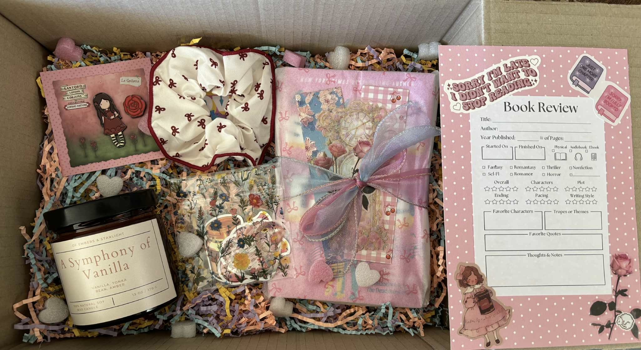 Book-themed gift box with a pink, romantic aesthetic, including a “Symphony of Vanilla” soy candle, a wrapped book tied with a sheer ribbon, a floral scrunchie, stickers and floral stationery, all arranged on pastel shredded paper next to a pink “Book Review” sheet decorated with cute illustrations and reading-related stickers.