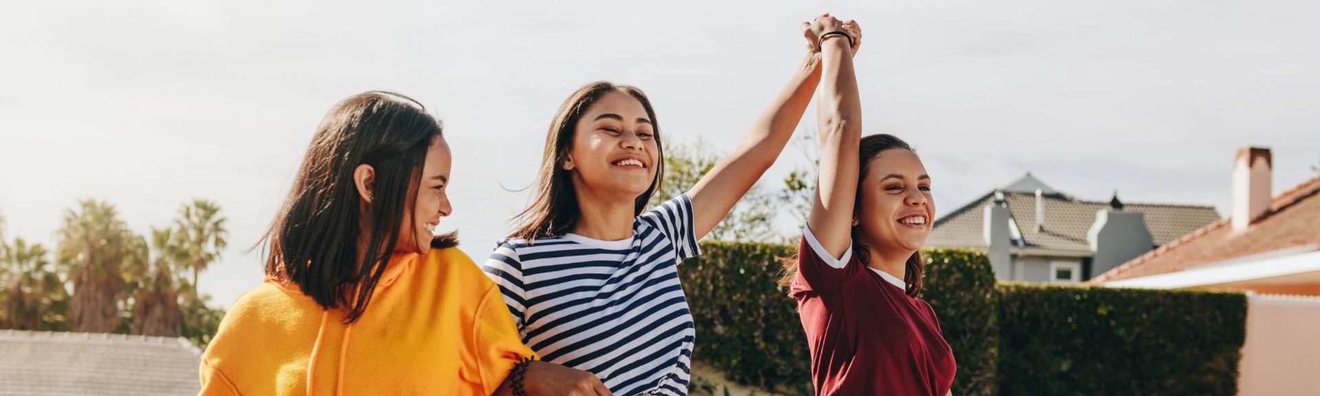 A vibrant outdoor scene featuring three young women smiling and celebrating under a bright sky. The leftmost woman wears a yellow hoodie, the middle woman a white t-shirt with blue stripes, and the rightmost woman a maroon t-shirt. Their raised arms and clasped hands convey joy and triumph, set against a backdrop of palm trees, green foliage, and suburban buildings.