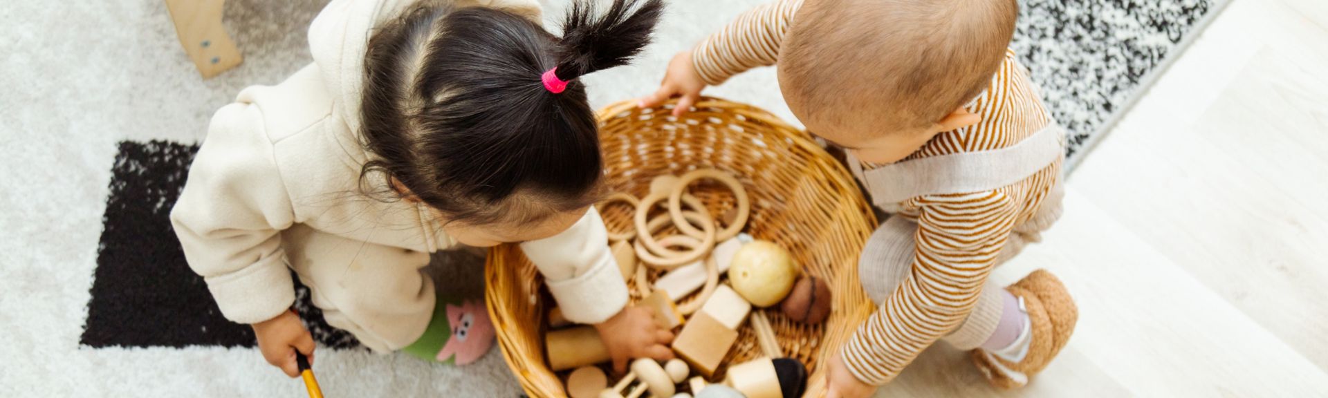 Two young children reaching into a wicker basket filled with wooden Montessori toys while tidying up together