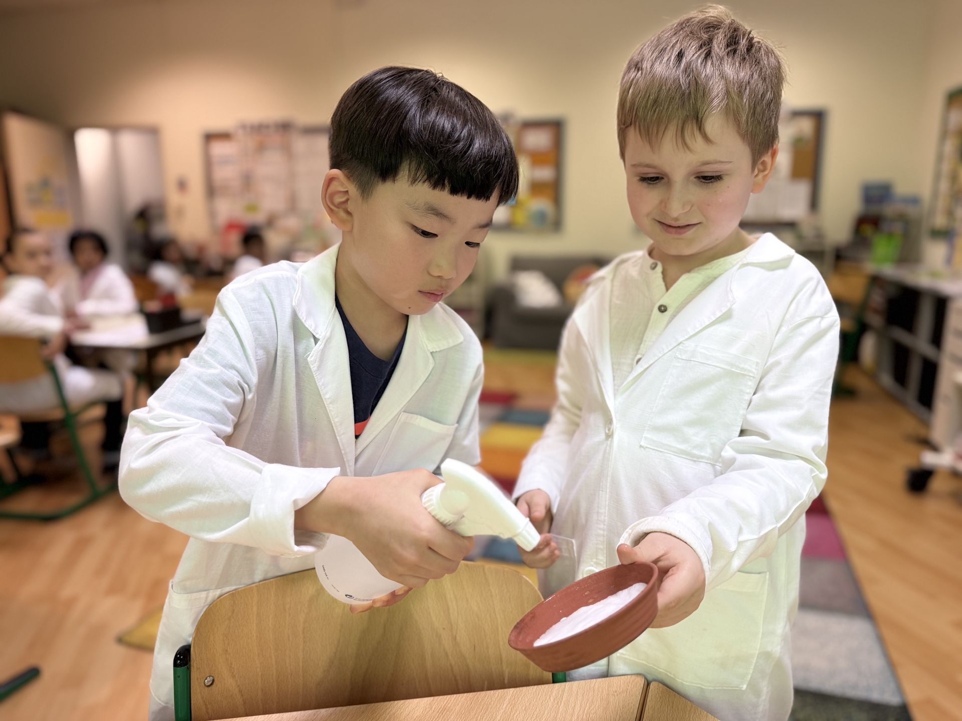 Two young students in white lab coats conduct a science experiment in a classroom, one holding a spray bottle while the other holds a small bowl of white foam or powder. Other students in lab coats are visible working at tables in the background.