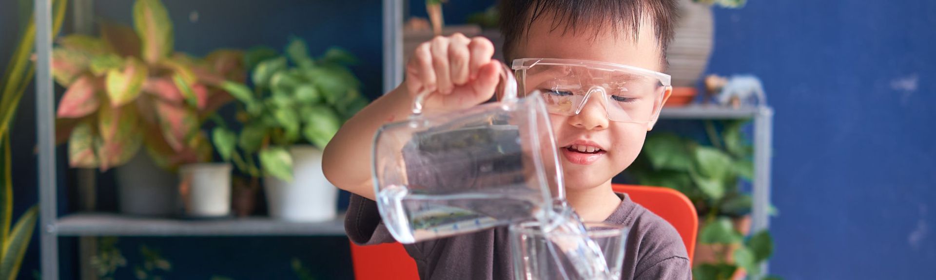 A young Asian boy wearing safety glasses is seated on a red chair, carefully pouring water from a clear plastic jug into a smaller container. He appears focused and curious. The background features blurred shelves with potted plants and a solid blue wall, creating a playful and educational atmosphere.