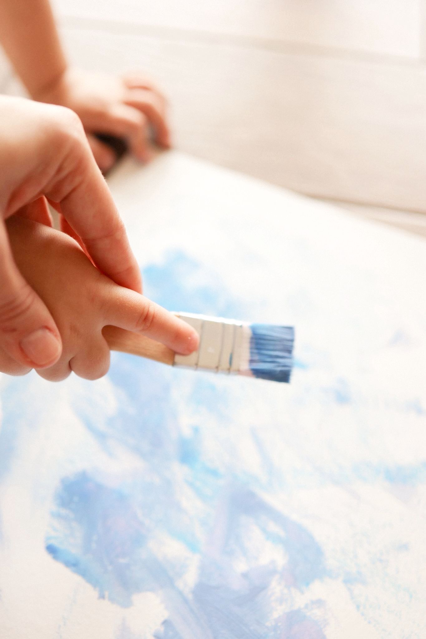 A close-up view of an adult's and a child's hands holding a paintbrush together, applying vibrant blue paint onto a white canvas. The canvas features abstract blue strokes, and the scene is brightly lit, emphasizing the collaborative and creative act of painting.
