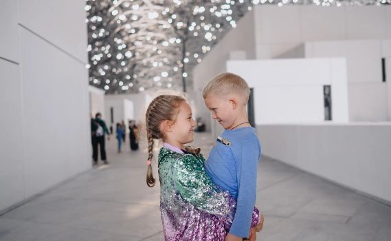 Two smiling children, a girl with braided hair and a shimmering jacket, and a boy in a blue shirt, share a warm moment in a modern building with a sparkling, star-like ceiling. The clean, minimalist interior and blurred figures in the background suggest a public cultural space.