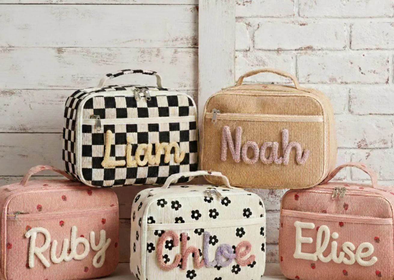 Five personalized soft-sided lunch boxes arranged on a rustic white wooden background. Each lunch box features a unique design and an embroidered name: 'Liam' on a black-and-white checkered pattern, 'Noah' on beige corduroy, 'Ruby' on pink corduroy with strawberry patterns, 'Chloe' on off-white corduroy with black floral patterns, and 'Elise' on pink corduroy with strawberry patterns.
