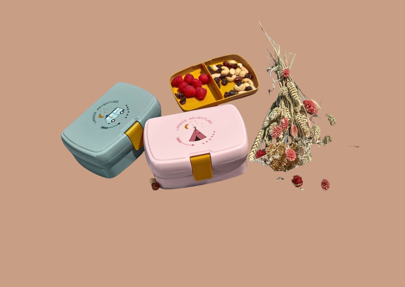 Two colorful bento lunchboxes with 'Classic Adventure' designs, one teal with a camper van graphic and one pink with a teepee graphic, placed on a beige background. A yellow snack tray with raspberries and a mix of cashews and raisins, along with a bouquet of dried flowers, completes the arrangement.