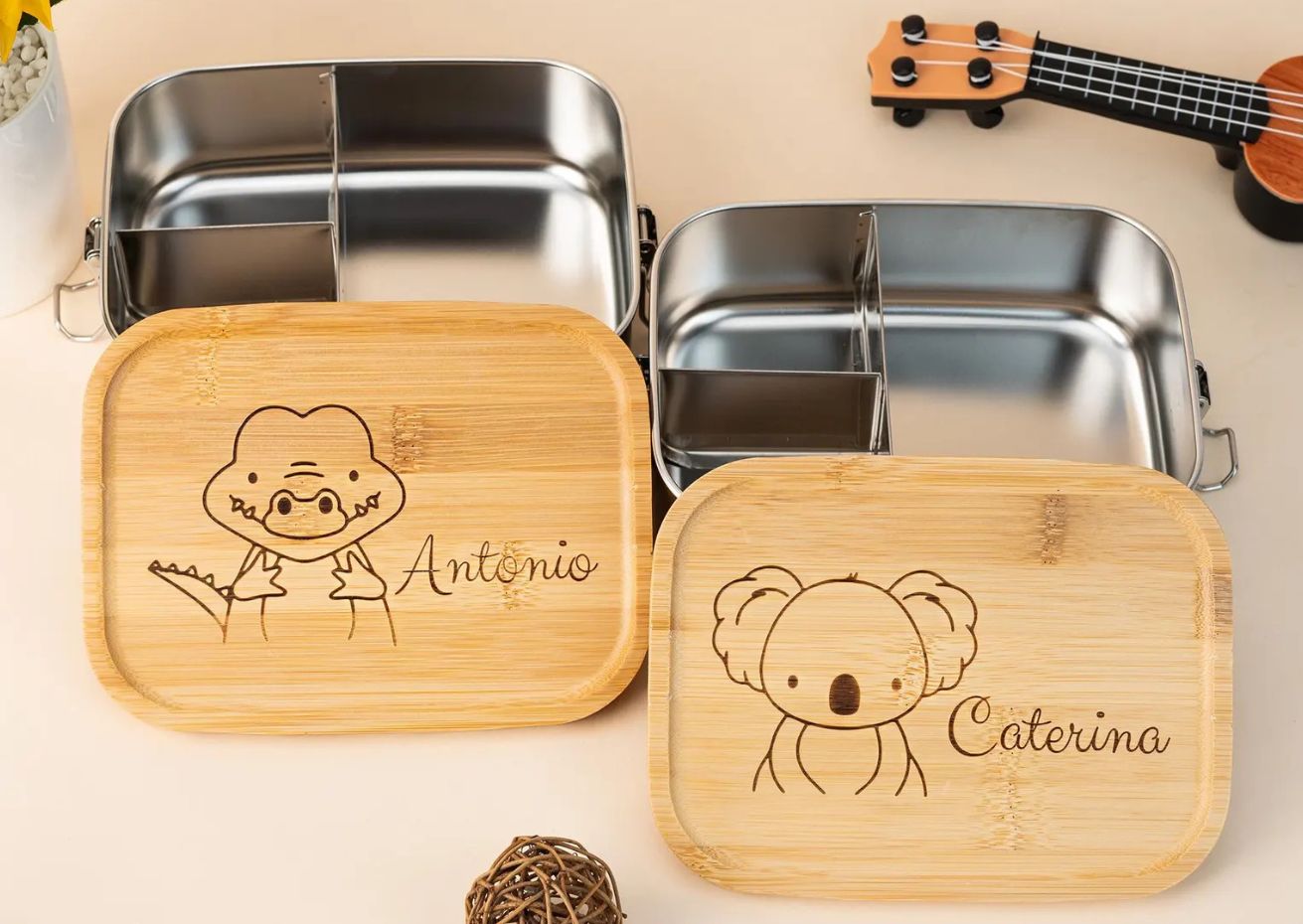 Two personalized stainless steel lunchboxes with bamboo lids, engraved with animal designs and names. The left lunchbox features an alligator and the name 'Antonio,' while the right one shows a koala and the name 'Caterina.' A small ukulele and a decorative wicker ball are also visible on a neutral background.