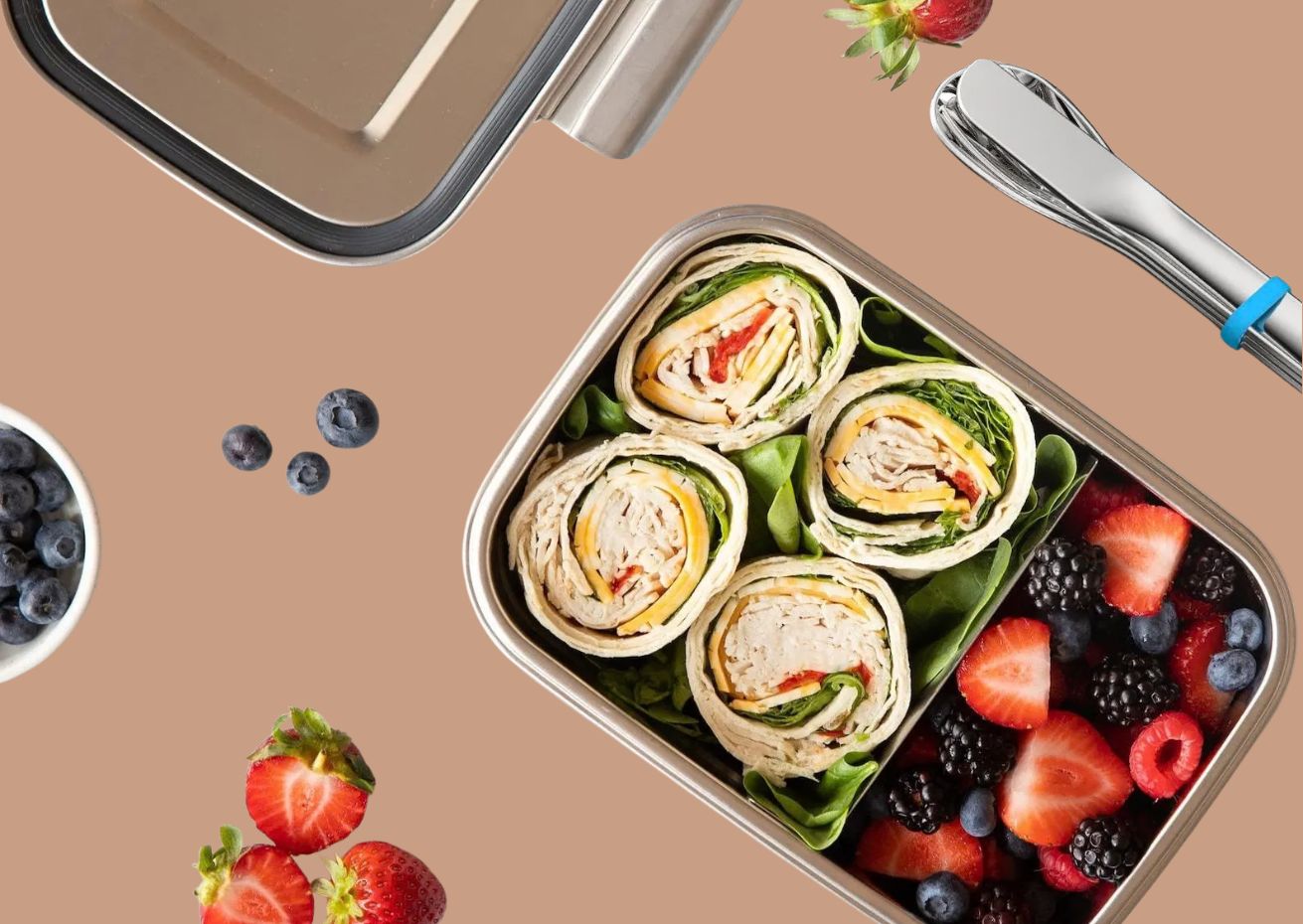 A stainless steel bento box with two compartments, one filled with pinwheel wraps on a bed of greens and the other with mixed berries. The lid, a set of stainless steel cutlery with a blue silicone band, and loose berries are arranged on a light brown background.