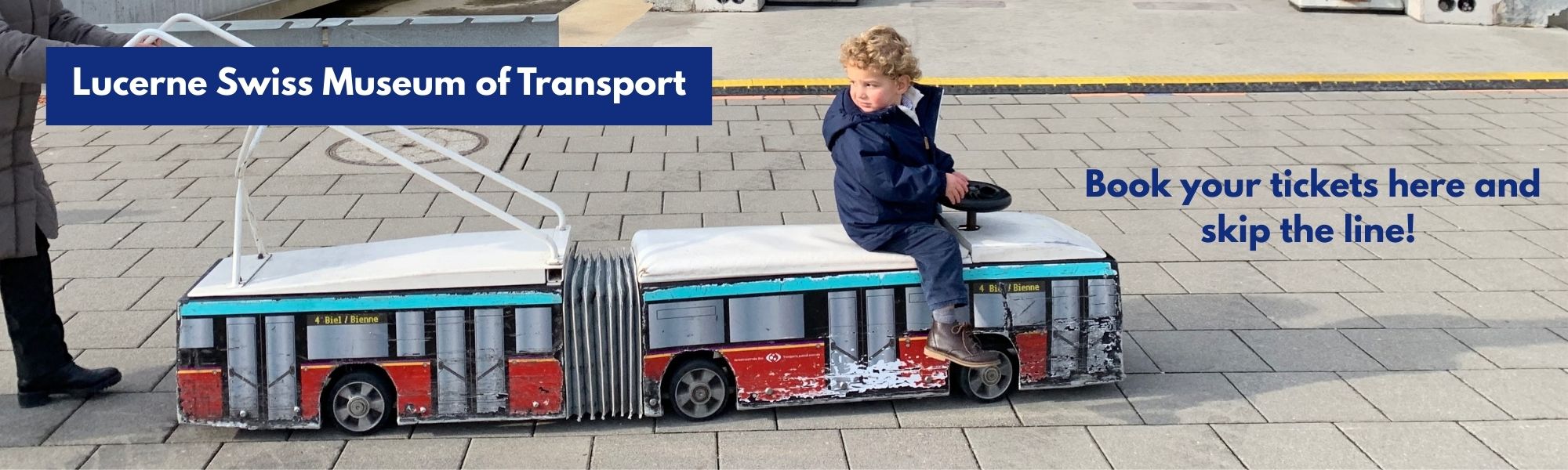 Image 2 Alt Text: A promotional banner for the Lucerne Swiss Museum of Transport showing a child with curly blonde hair sitting on a toy articulated bus or tram, being pushed by an adult. The toy vehicle is red, blue, and white, with "4 Biel / Bienne" text on its side. The scene is set on a paved surface with a light concrete structure and a yellow line in the background. The text "Lucerne Swiss Museum of Transport" and "Book your tickets here and skip the line!" is prominently displayed.