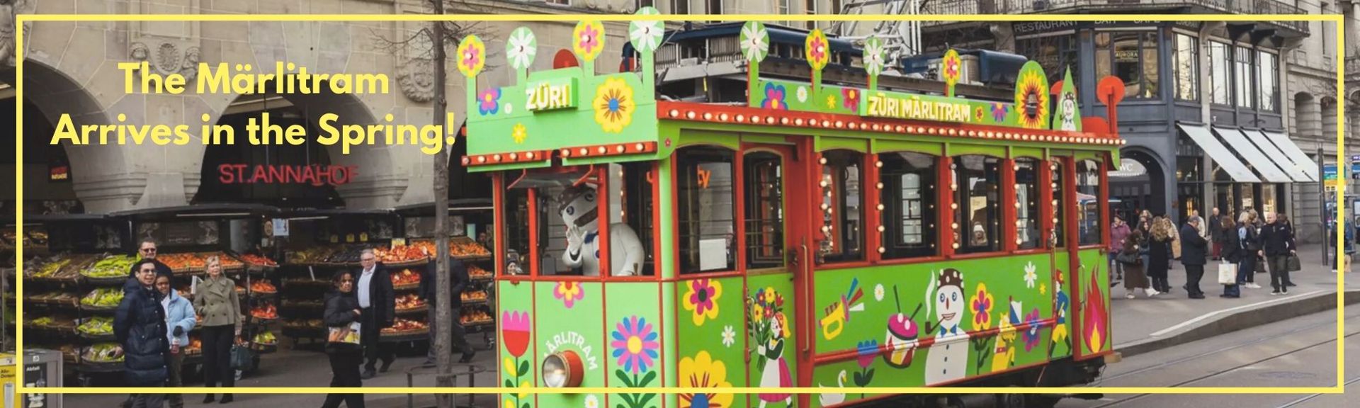 A vibrant green Märlitram tram decorated with colorful flowers, stars, and whimsical illustrations, including a snowman and a girl holding flowers, on a Zurich street. The tram features the text 'ZÜRI MÄRLITRAM' and 'MÄRLITRAM' on its exterior. A yellow overlay at the top reads 'Das Märlitram fährt jetzt auch im Frühling!' in German. The background includes the St. Annahof building, a fruit stall, and people walking nearby.
