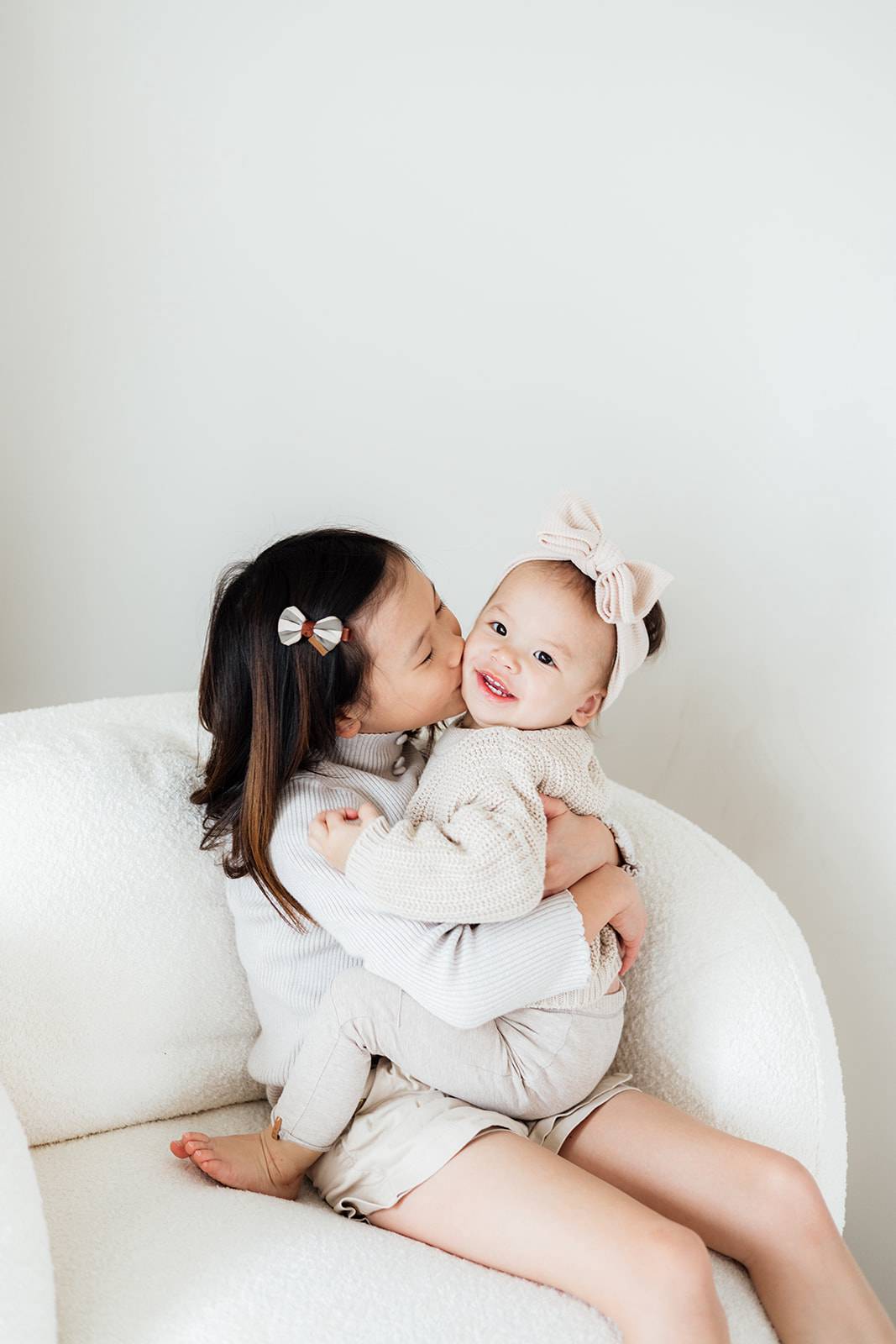 A sweet kiss of one sister to her baby sister captured by Mary Fernandez family photographer in Switzerland.  The children are asian and photographed in a white arm chair with a white background.  