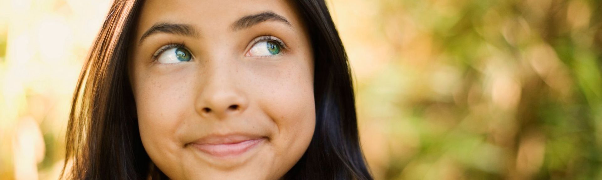 Close-up portrait of a young child outdoors, looking to the side with a gentle smile, soft natural light and greenery in the background, conveying curiosity and calm.