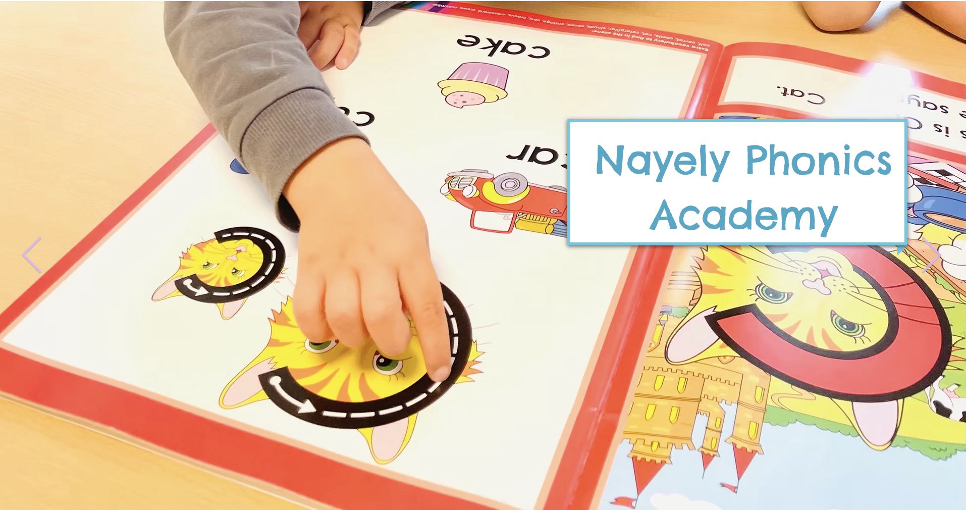 An open word and picture book for children 4-8 learning english.  There is a young child's hand pointing at a cat picture in the book, during class reading and pointing at Nayeli Phonics Academy in Zollikon.  The logo in white and blue is on the right hand side.  