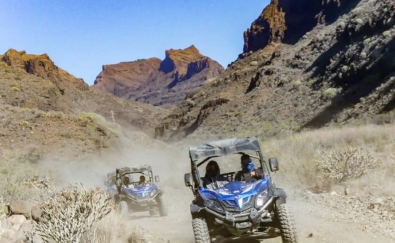 Two blue dune buggies driving on a dusty, unpaved road through a rugged, arid mountain landscape under a clear blue sky. The lead buggy, with two helmeted occupants, kicks up a trail of dust, while the second buggy follows closely behind. The rocky terrain and sparse vegetation emphasize the adventurous off-road setting.