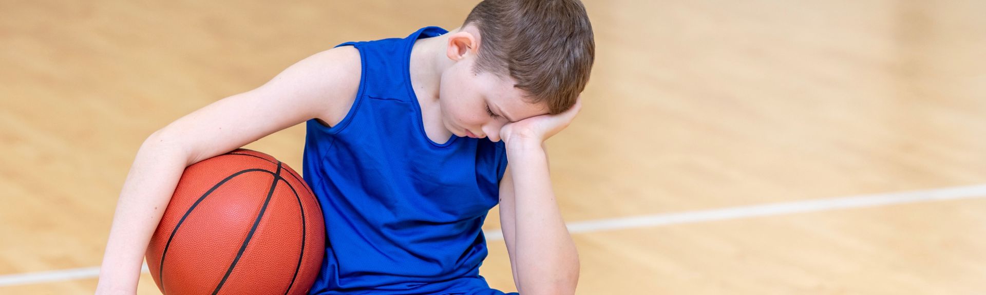 A teenage boy sits on a basketball court, wearing a bright blue sleeveless athletic jersey. He looks down with his head resting on his right hand, appearing sad or disappointed. His left arm holds an orange basketball against his side. The wooden court floor and a white line in the background suggest an indoor setting. The image conveys a sense of melancholy or exhaustion related to sports.