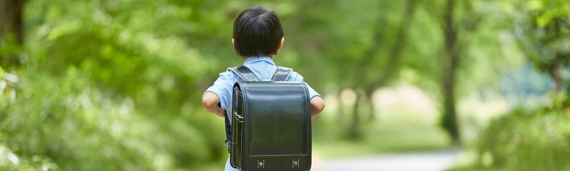 A young child with short dark hair walks down a lush green path, wearing a light blue shirt and a dark green school backpack with white stitching. The background features blurred greenery, creating a serene and natural outdoor setting.