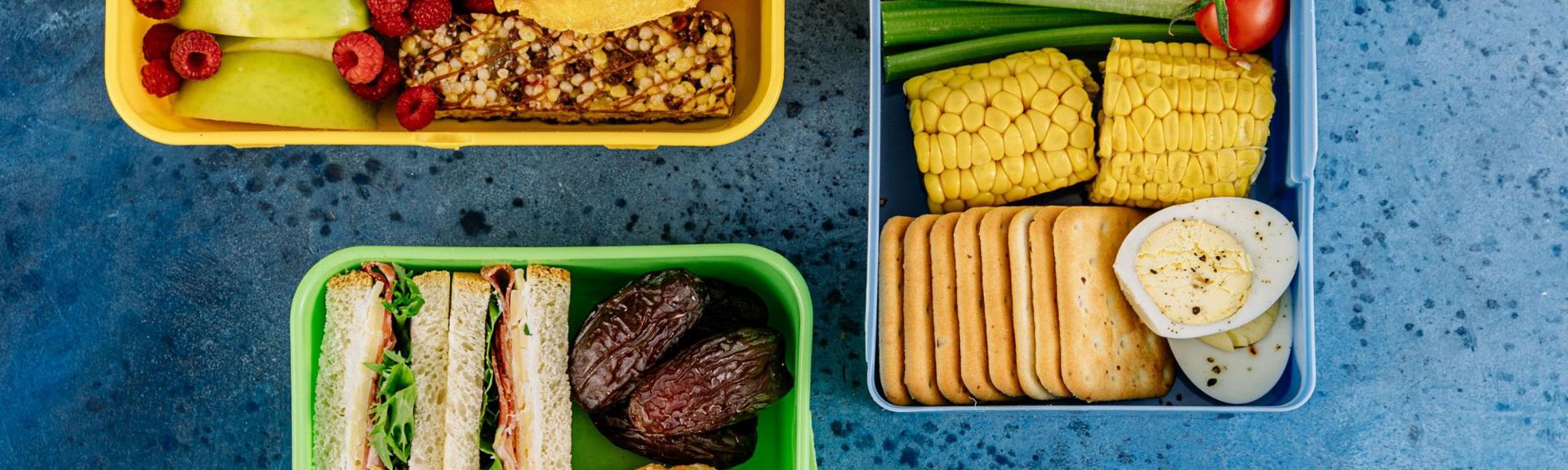 er Alt text: "Overhead view of three colorful lunchboxes on a blue textured surface, each filled with a variety of fresh and healthy foods. The yellow lunchbox contains sliced green apples, red raspberries, and a granola bar. The blue lunchbox features celery sticks, a cherry tomato, corn on the cob, crackers, and hard-boiled egg slices. The green lunchbox holds small sandwiches with leafy greens, meat, and cheese, alongside plump brown dates.