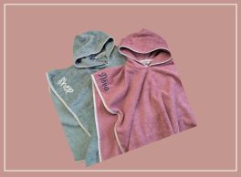Two personalized hooded towel ponchos displayed on a muted pink background. The left poncho is greenish-gray with 'Shep' embroidered in white, and the right poncho is pink with 'Nove' embroidered in maroon. Both feature white trim and are made of soft, towel-like material, showcasing their cozy and customizable design.