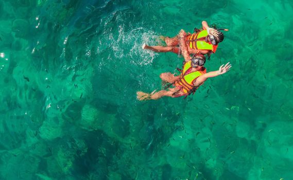 An aerial view of two snorkelers in bright yellow-orange life vests swimming in clear, turquoise-green tropical waters. The sunlight illuminates the rippling surface, revealing patches of varying depths and small fish, creating a serene aquatic scene.