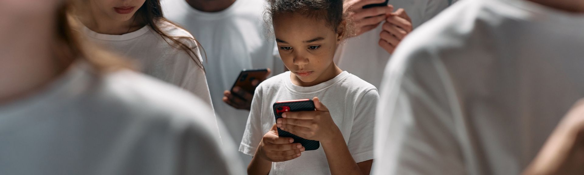 Young girl focused on her smartphone while surrounded by adults, illustrating children's screen time and social media use.