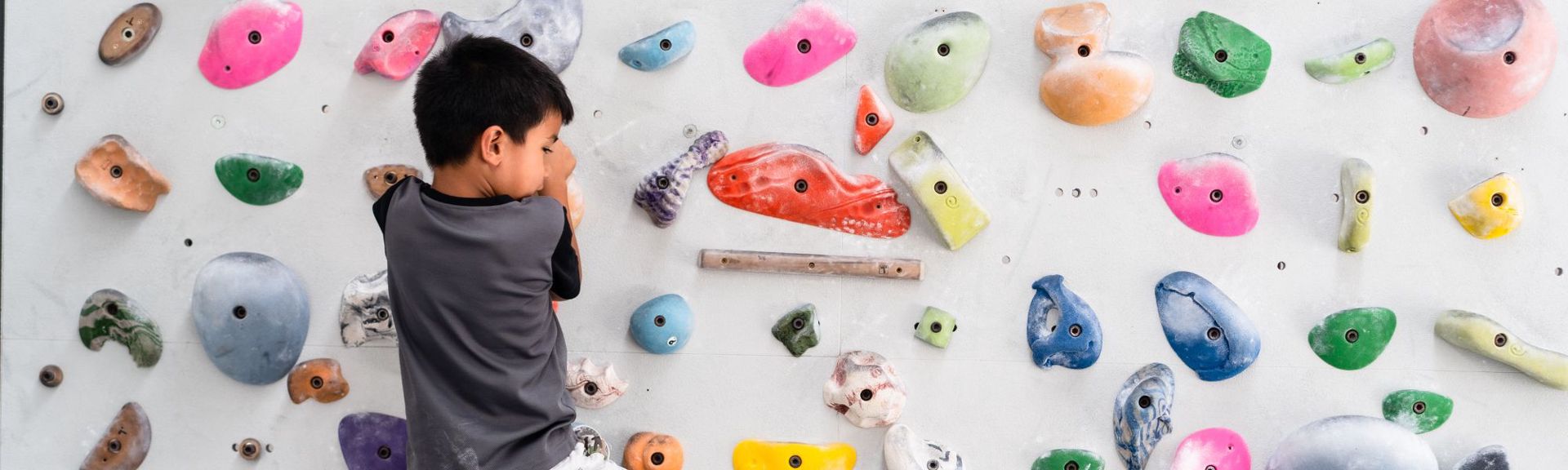 A young child bouldering on an indoor climbing wall filled with colorful holds of various shapes and sizes. The child, wearing a dark grey and black t-shirt, is gripping an orange climbing hold while looking upward, surrounded by a vibrant array of climbing routes. The wall features visible chalk marks and empty bolt holes, indicating a customizable climbing setup.