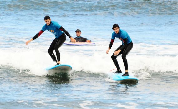 Three individuals in the ocean during a surfing session. Two surfers, wearing bright blue and black wetsuits, are standing on light blue surfboards, balancing on small waves with water splashing around them. A third person, partially submerged in the water and holding a yellow bodyboard, observes or assists. The scene captures a fun and dynamic surfing activity in clear, sunlit ocean waters.