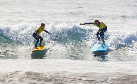 Two surfers riding ocean waves under bright sunlight. The surfer on the left is on a yellow surfboard, crouched with arms extended for balance, surrounded by white foam from the breaking wave. The surfer on the right is on a blue surfboard in a similar stance. Both wear dark wetsuits with bright yellow accents. The scene captures the energy of surfing amidst greenish-blue waves and sparkling water.