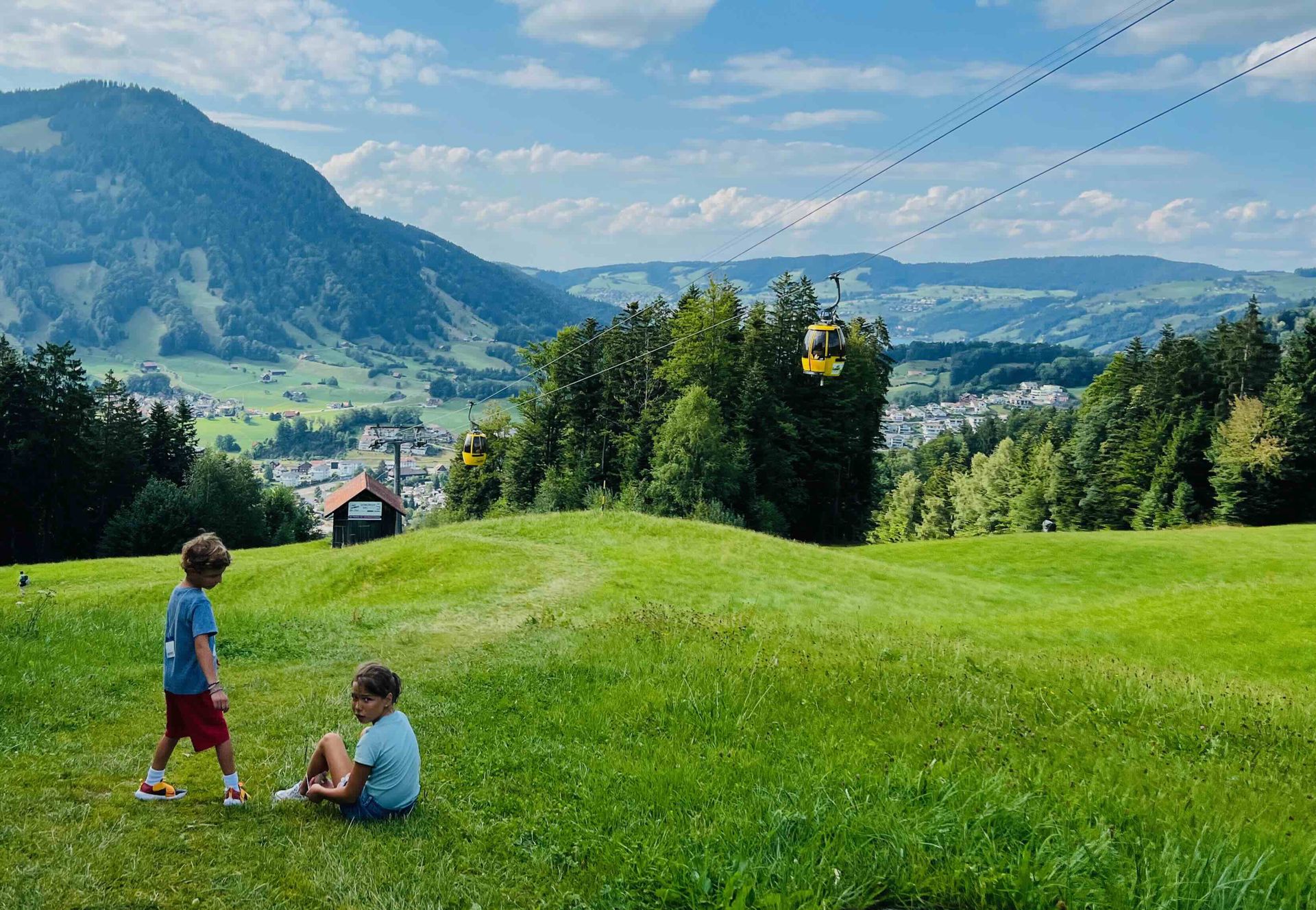 Two children rest on a grassy hillside in the foreground of a picturesque alpine landscape. A boy stands wearing a blue t-shirt and red shorts, while a girl sits nearby in a light blue t-shirt and denim shorts, looking back toward the camera. In the midground, a rustic wooden hut and two yellow gondola cabins suspended on cables are visible, set against a backdrop of dense green trees. The background features a valley with a small village and rolling mountains under a bright blue sky with scattered white clouds, evoking a serene summer day in the Alps.