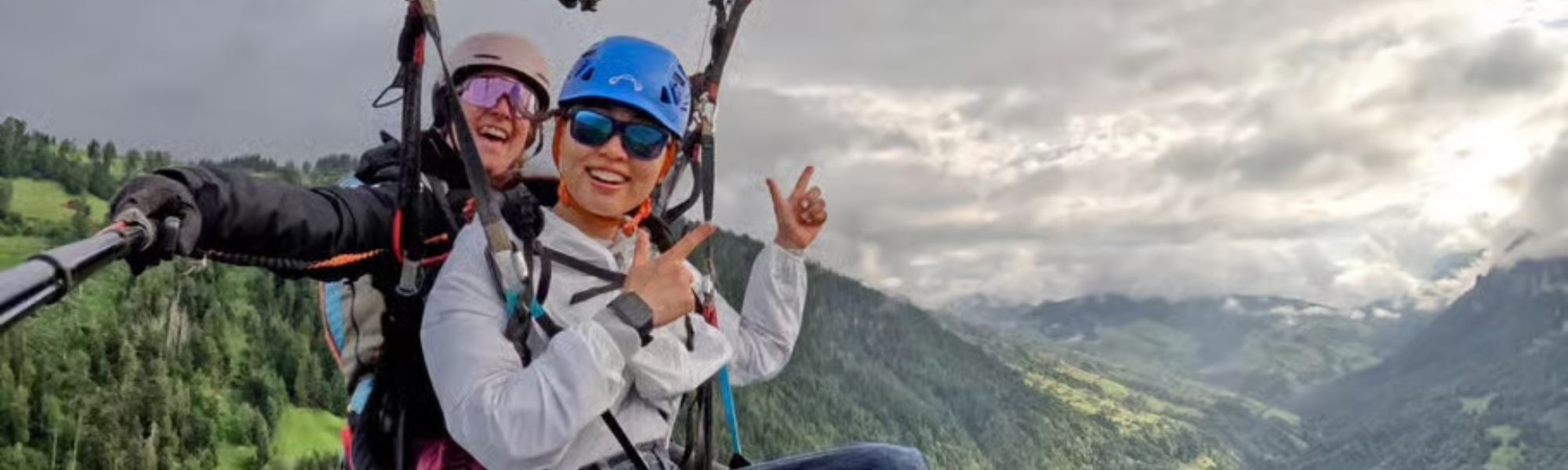 A tandem paragliding flight over a lush green valley and forested mountains. The passenger in the foreground, wearing a bright blue helmet, white jacket, and blue-tinted sunglasses, smiles and gestures toward the camera. The pilot behind, in a tan helmet and iridescent goggles, holds a selfie stick to capture the moment. The background features rolling green hills, misty mountain peaks, and an overcast sky, creating a dramatic and adventurous scene.