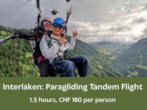 Two people enjoy a tandem paragliding flight over a lush, mountainous valley in Interlaken. The passenger in the front wears a blue helmet, dark sunglasses, a white jacket, and blue jeans, giving a thumbs-up and peace sign. The pilot behind wears a beige helmet and purple-tinted goggles, smiling while holding a selfie stick. The background features steep green slopes descending into a deep valley under an overcast sky, creating a thrilling and scenic adventure atmosphere.