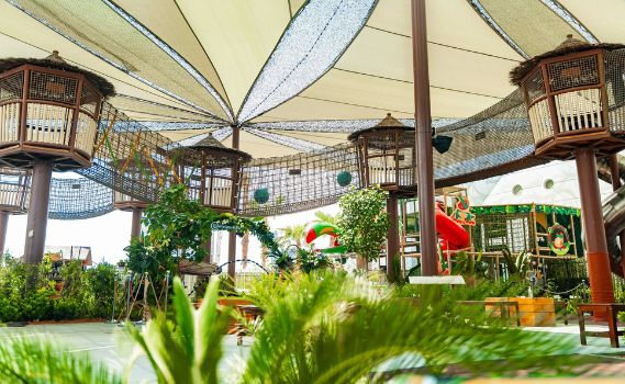 Jasper "An outdoor adventure play area featuring elevated wooden structures resembling treehouses, connected by rope bridges under a large beige canopy. The scene is surrounded by lush green foliage, with a red slide and other colorful play equipment visible. The vibrant and shaded environment is designed for family-friendly fun and exploration.