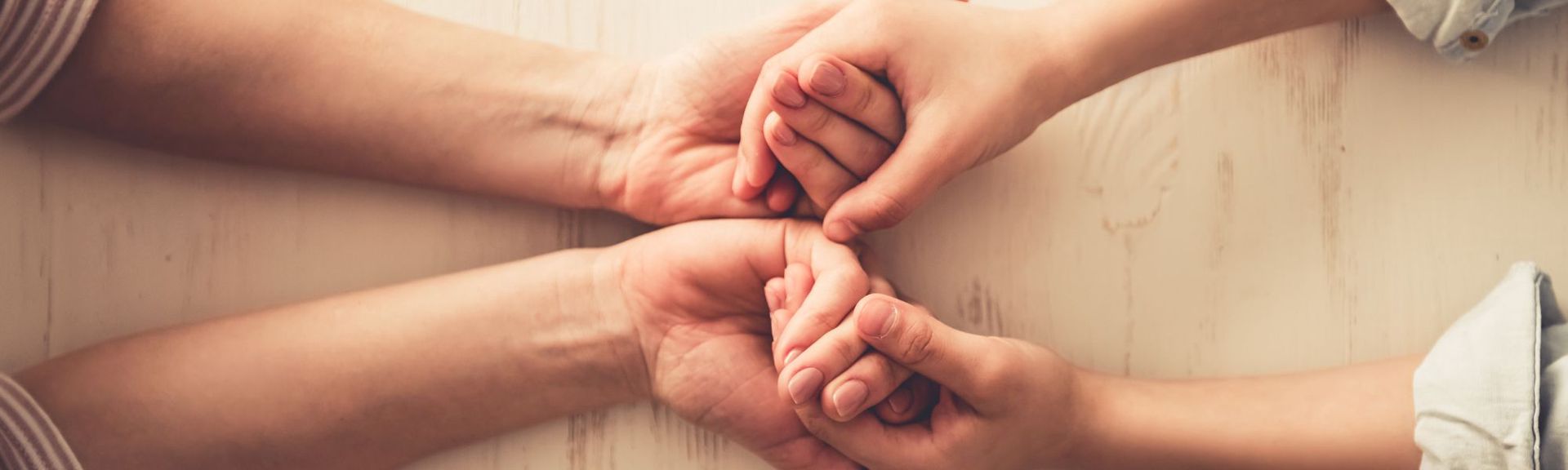 A close-up, top-down view of an adult's hands gently holding a child's hands on a light wooden surface. The adult's hands, showing subtle signs of maturity, cradle the smaller, delicate hands of the child, symbolizing care and responsibility. The soft lighting and natural tones evoke warmth and intimacy, reflecting the emotional and physical support mothers provide while carrying the unseen mental load of nurturing, protecting, and guiding their children.