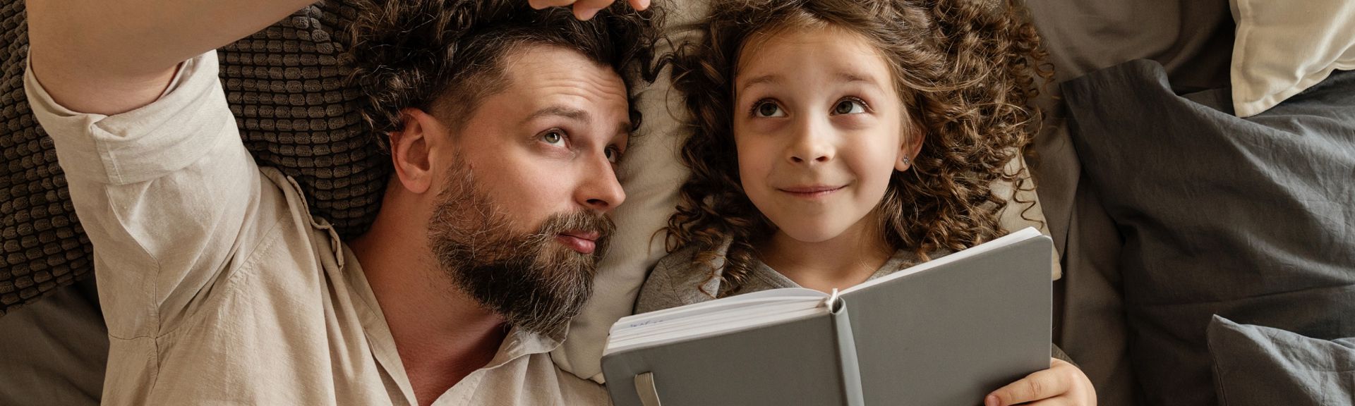 Father and daughter lying down together reading a book, sharing a cozy low-energy moment on the couch