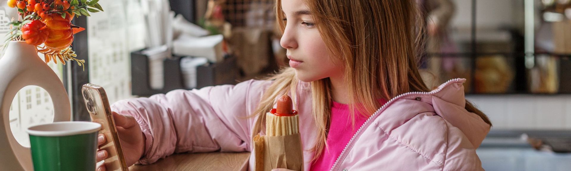  young girl in a light pink puffy jacket and fuchsia top is seated in a cozy cafe, holding a hotdog with ketchup in her left hand and a smartphone in her right. Her light brown, golden-highlighted hair falls over her shoulders as she looks intently at her phone. A green paper cup and a white vase with autumn-themed decorations, including orange berries and mini pumpkins, sit on the table in front of her. The softly blurred background suggests a warm, casual atmosphere.