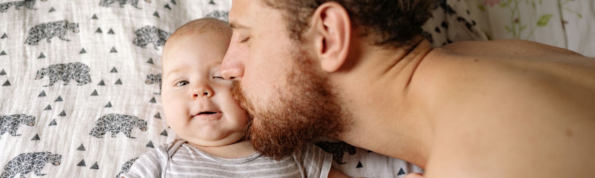 Father tenderly kissing his baby on the cheek while lying on a patterned blanket, symbolizing the deep emotional connection and transformative journey of parenthood.