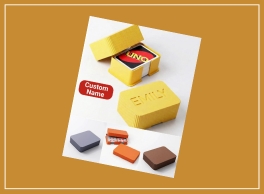 A product display featuring customizable card game cases on a white sheet against an orange-brown background. The central focus is a yellow case, partially open, revealing a deck of UNO cards, with another closed yellow case embossed with "BMY" beside it. A red label reading "Custom Name" points to the yellow cases, highlighting personalization options. Below, four smaller square cases in gray-blue, orange-red, light orange, and brown are arranged horizontally, showcasing additional color options or product variations. The image emphasizes customizable storage solutions for card games.