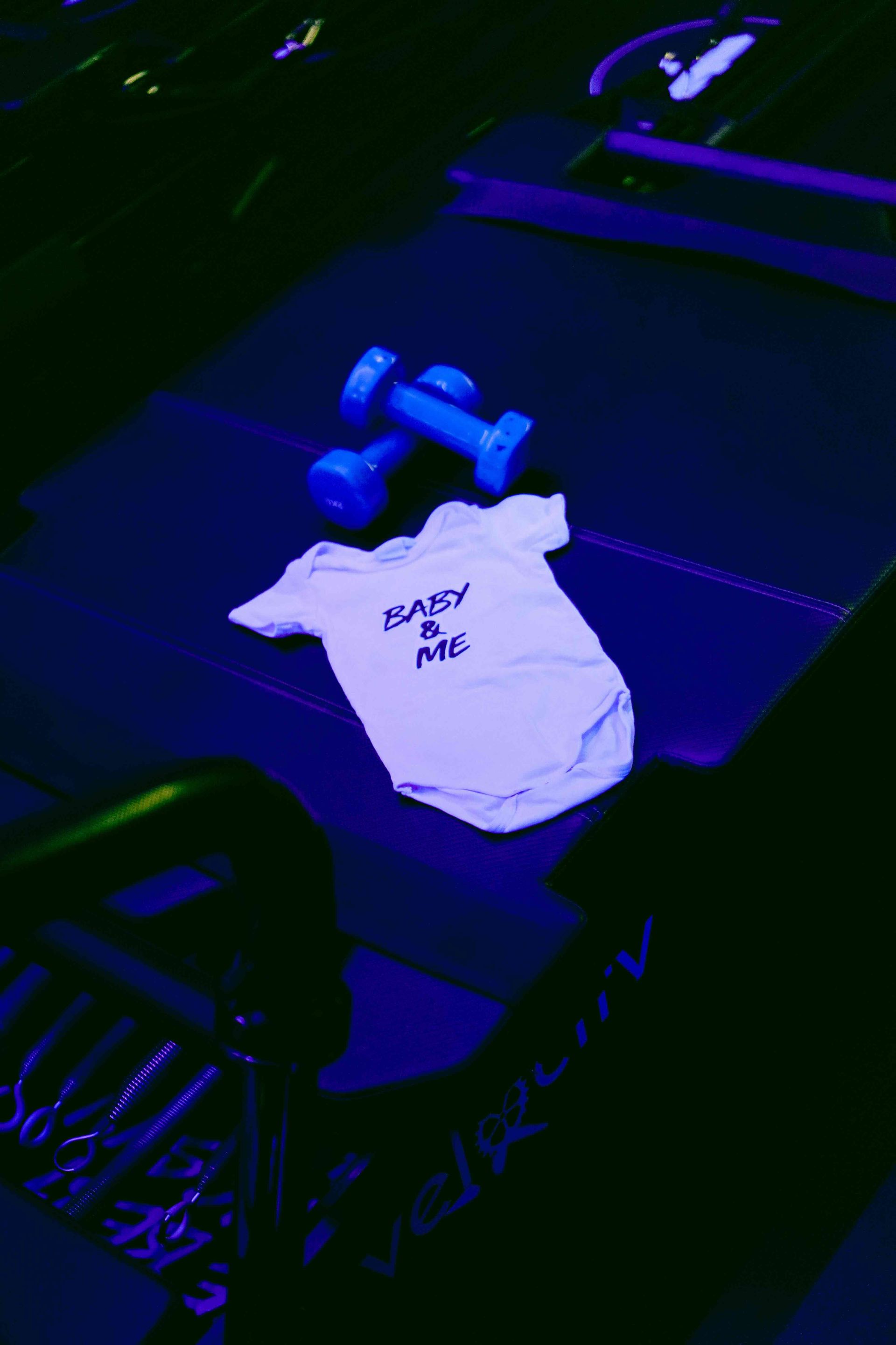 A white baby onesie with the text "BABY & ME" is laid flat on a dark surface, surrounded by two blue dumbbells. The setting features dim, atmospheric lighting with blue, purple, and green hues, suggesting a fitness studio. Parts of exercise equipment, including metal springs and a bar, are visible in the foreground, while the word "VELOCITY" is partially visible on the lower right. The image conveys a theme of postnatal fitness or parent-and-baby workout classes.