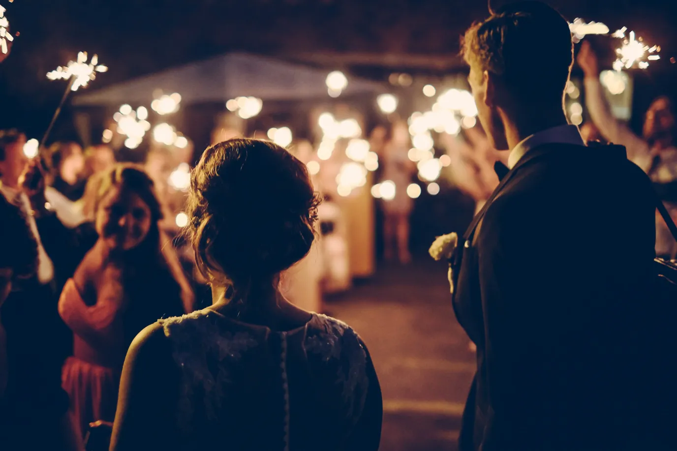 A nighttime wedding reception scene viewed from behind a couple. In the foreground, the back of a woman in a dark dress and a man in a suit are visible. They are facing a crowd of people holding sparklers, creating a warm, glowing atmosphere. A building with string lights is visible in the background, adding to the festive ambiance.
